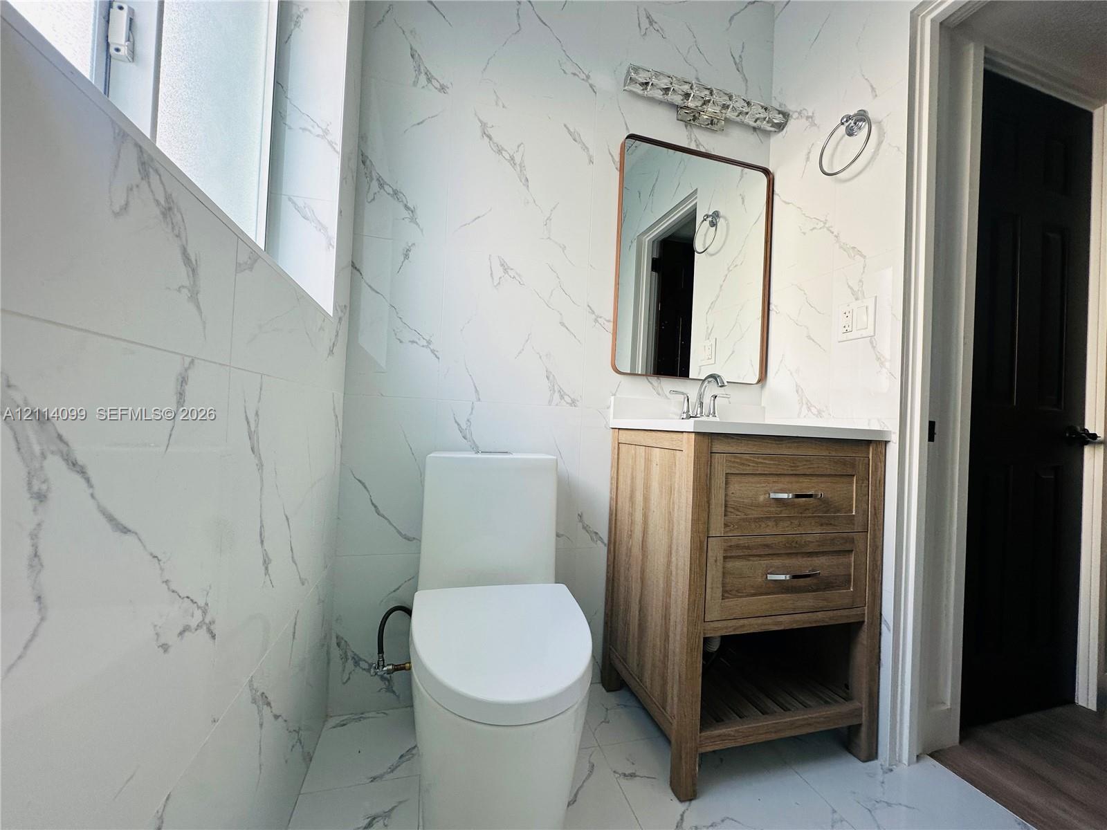 Bathroom featuring floor-to-ceiling stone-finish wall tile, wood-finish vanity with a rectangular sink, wall-mounted rectangular mirror, and modern elongated toilet