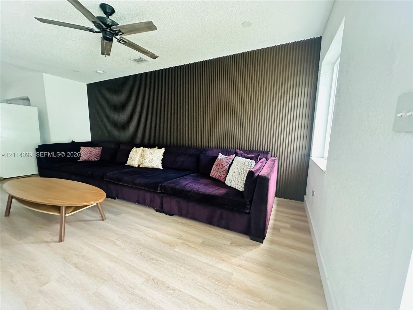 Accent wall with vertical fluted paneling, light-tone wood-finish flooring, and a multi-blade ceiling fan
