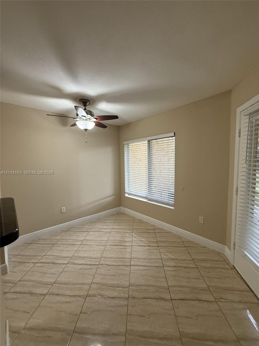 Neutral-toned interior featuring large-format tile flooring, a central ceiling fan with integrated lighting, horizontal window blinds, and a glass-paneled exterior door