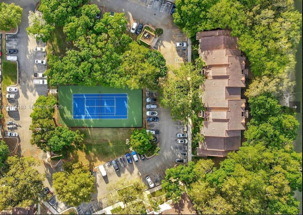 Aerial perspective of the property grounds featuring a multi-level building with shingled roofing, a tennis court, designated parking areas, and mature tree canopy