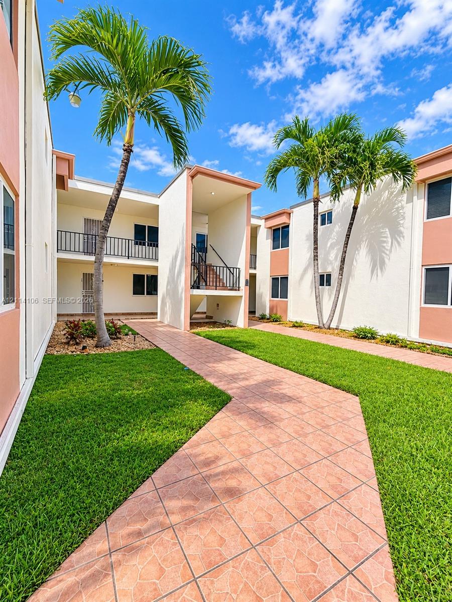 Exterior courtyard featuring a terracotta-tiled walkway, manicured lawn, palm trees, and multi-level buildings with metal railings
