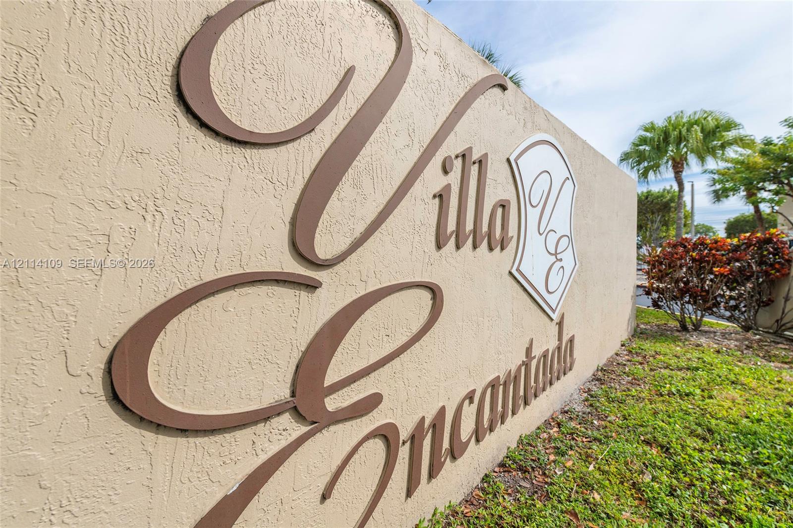 Textured stucco monument signage featuring dimensional metal lettering and a decorative emblem, accented by professional landscaping with palm trees and shrubbery