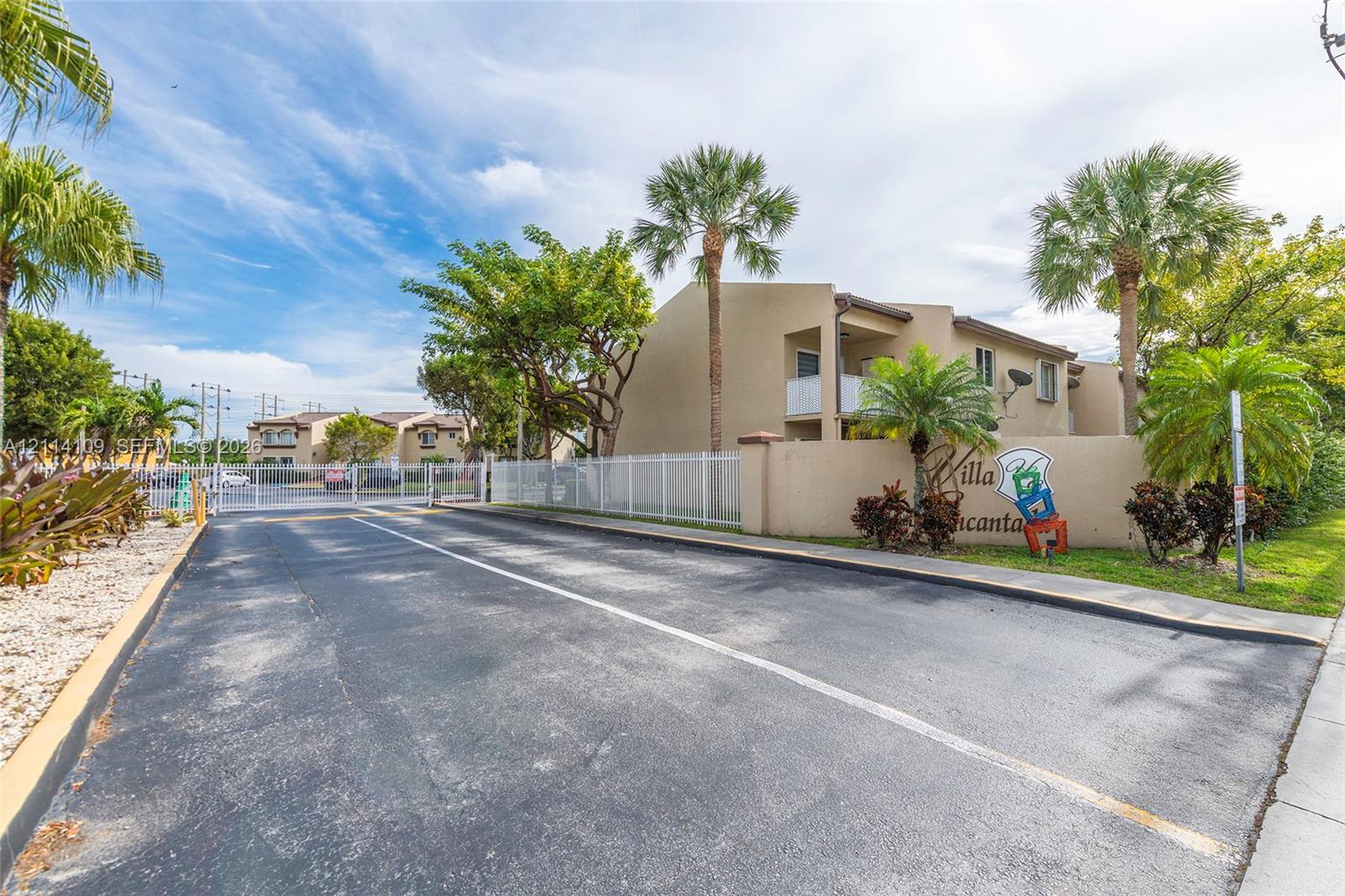 Gated entrance featuring stucco exterior walls, paved driveway, white perimeter fencing, and mature palm landscaping