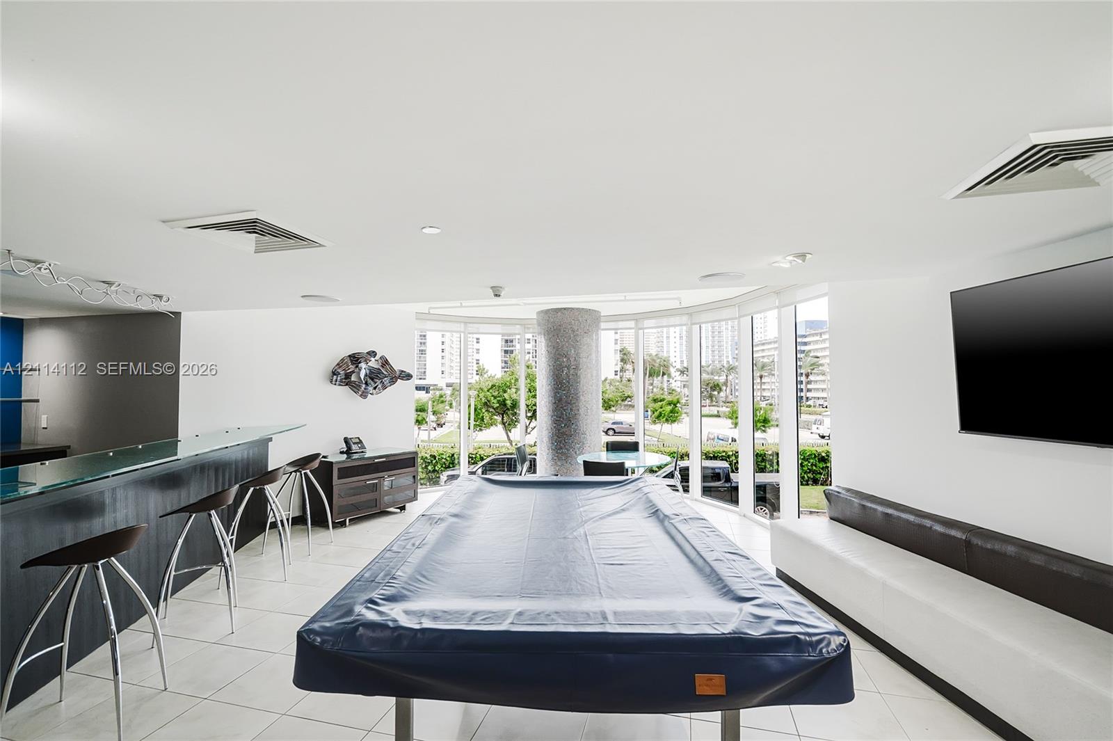Expansive recreation area featuring floor-to-ceiling glass walls, a built-in wet bar with glass countertop, tile flooring, and a stone-clad structural column