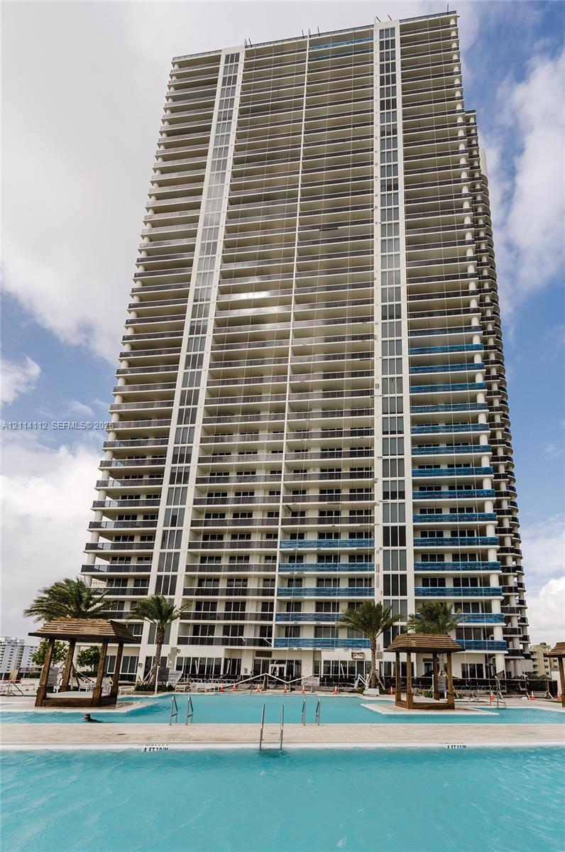 High-rise condominium tower featuring glass balcony railings, extensive fenestration, and a multi-section swimming pool with integrated sun decks and wooden cabanas