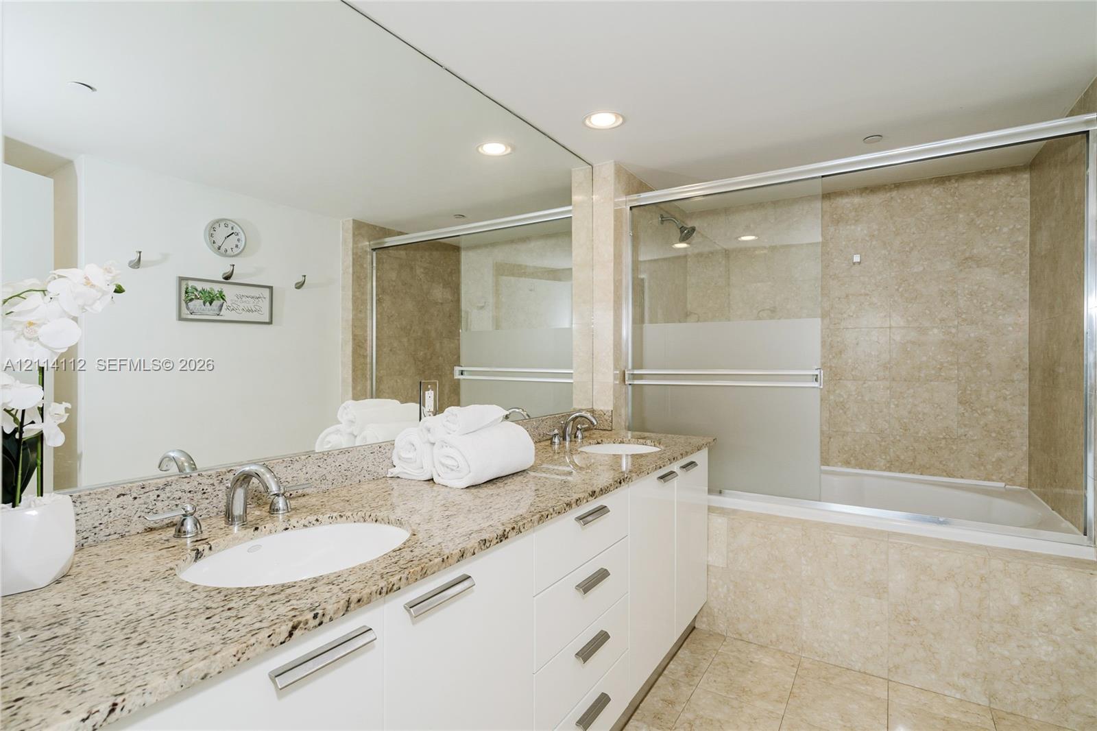 Full-width vanity with stone countertop, dual undermount sinks, contemporary white cabinetry, and tub-shower combination featuring sliding glass doors and tiled surround