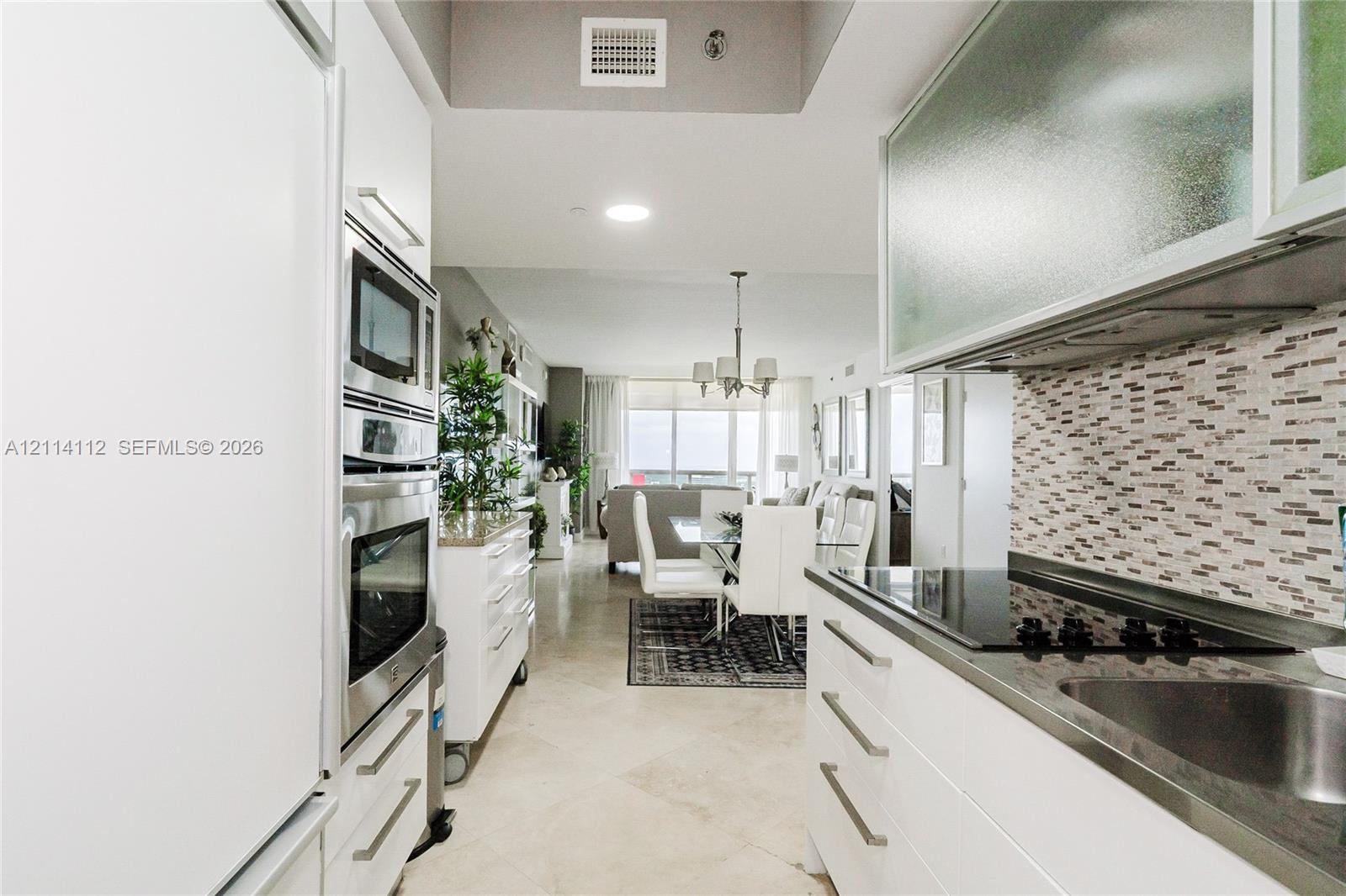 Galley kitchen featuring white flat-panel cabinetry, stainless steel wall-mounted oven and microwave, mosaic tile backsplash, and stone flooring