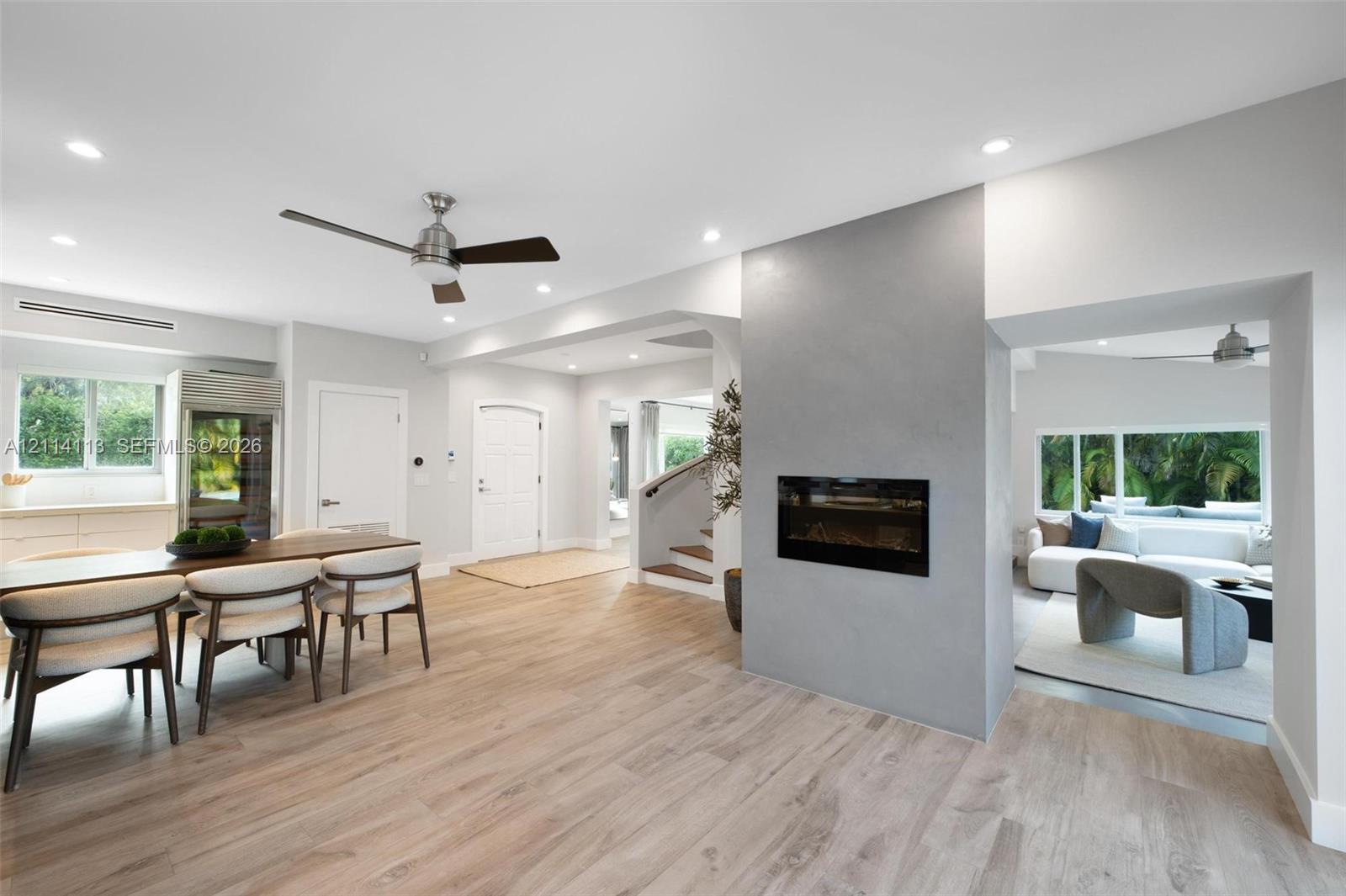 Light-toned wood-finish flooring, central fireplace with a grey smooth-finish surround, recessed ceiling lighting, ceiling fans, and multi-paned windows