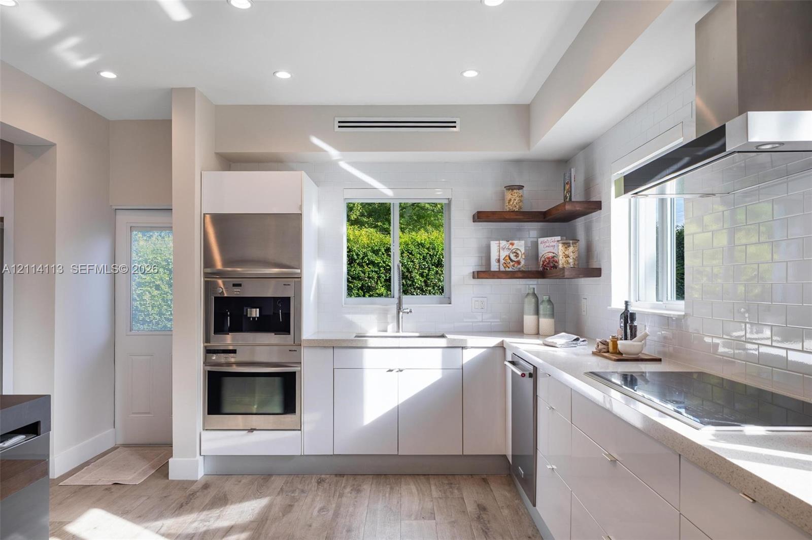 Kitchen featuring white cabinetry, wood-finish flooring, subway tile backsplash, stainless steel built-in appliances, and floating wood shelving