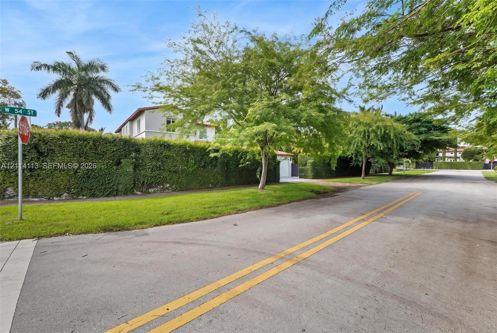 View of the side W 54th street featuring a tall perimeter privacy hedge, manicured lawn, and mature trees