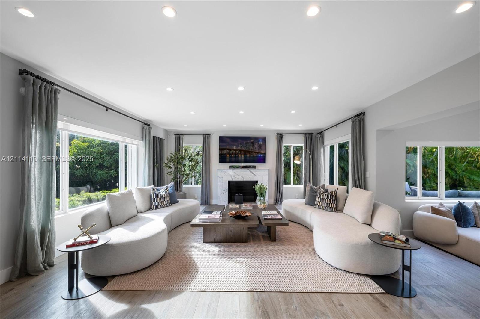 Living area featuring wood-finish flooring, recessed lighting, expansive multi-pane windows, and a central fireplace with a stone-finish surround