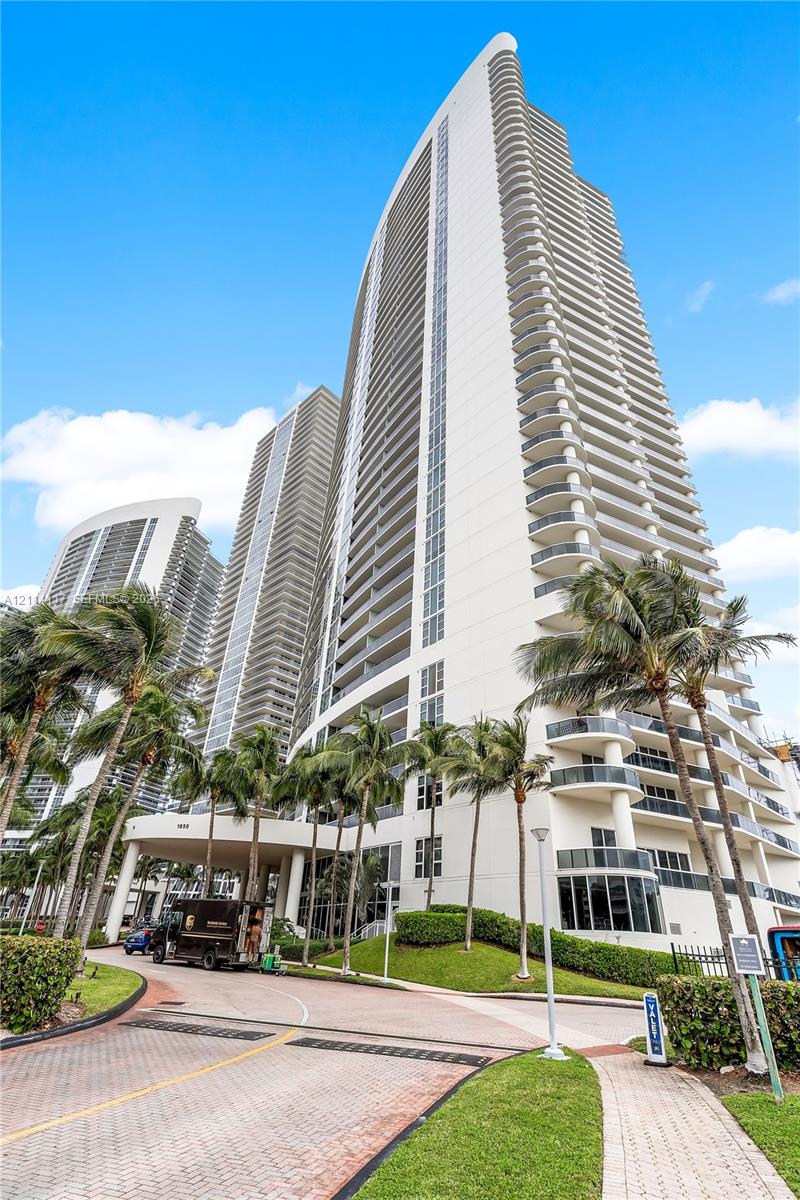 High-rise tower featuring a curved architectural facade, extensive private balconies, a covered porte-cochre entry, and surrounding tropical landscaping