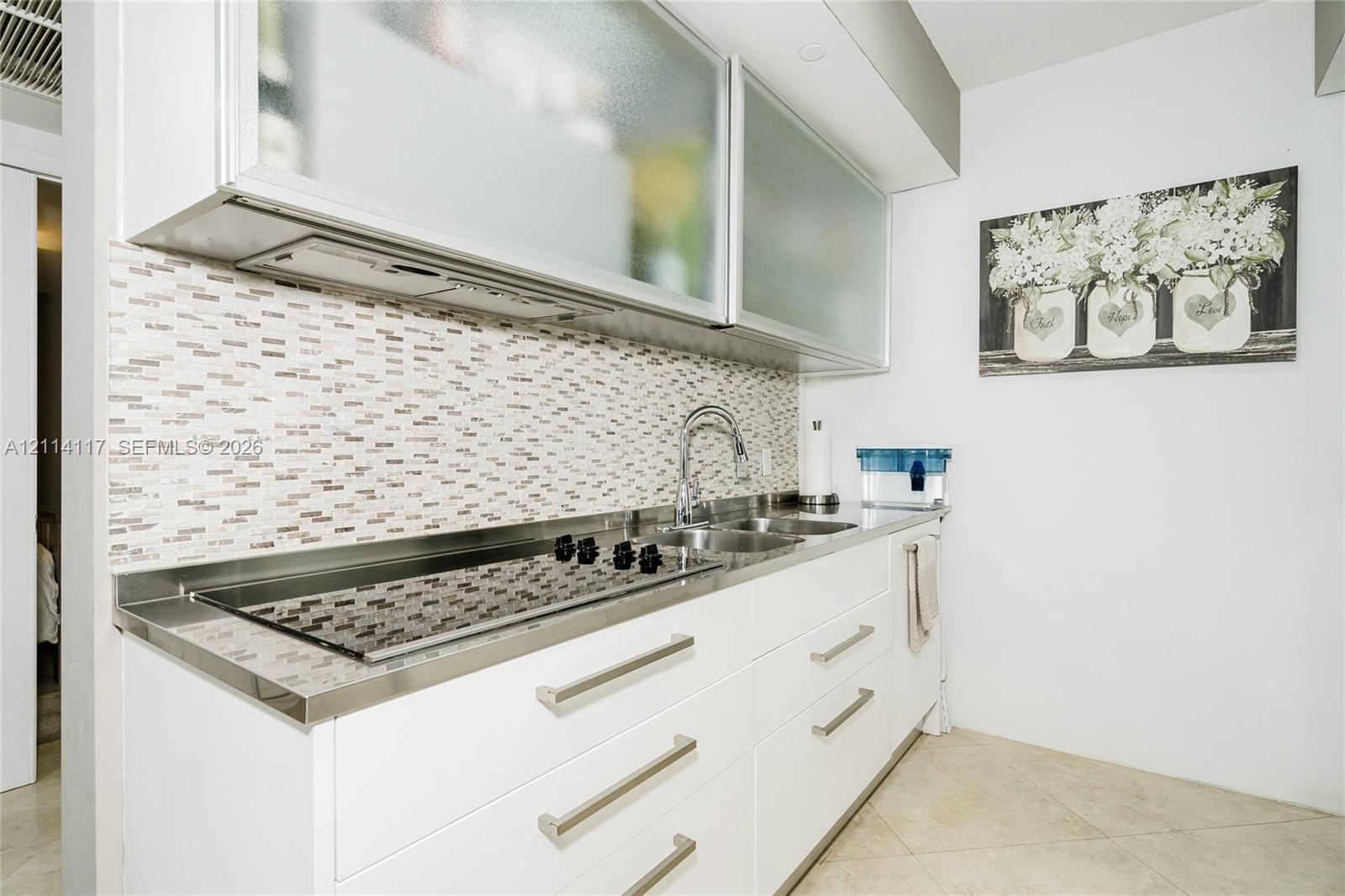 Kitchen featuring white flat-panel cabinetry, frosted glass upper cabinet doors, mosaic tile backsplash, integrated stainless steel cooktop, and metal hardware