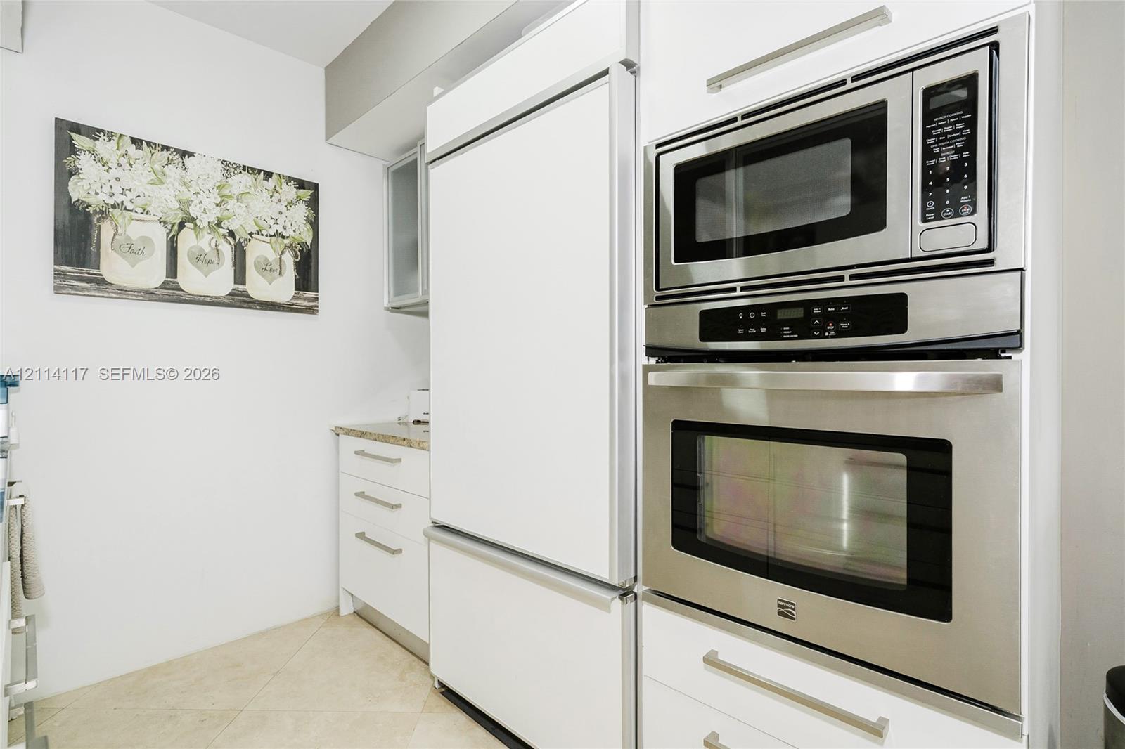 Stainless steel built-in microwave and wall oven, integrated panel-ready refrigerator, white shaker-style cabinetry with satin nickel hardware, and stone-finish countertops