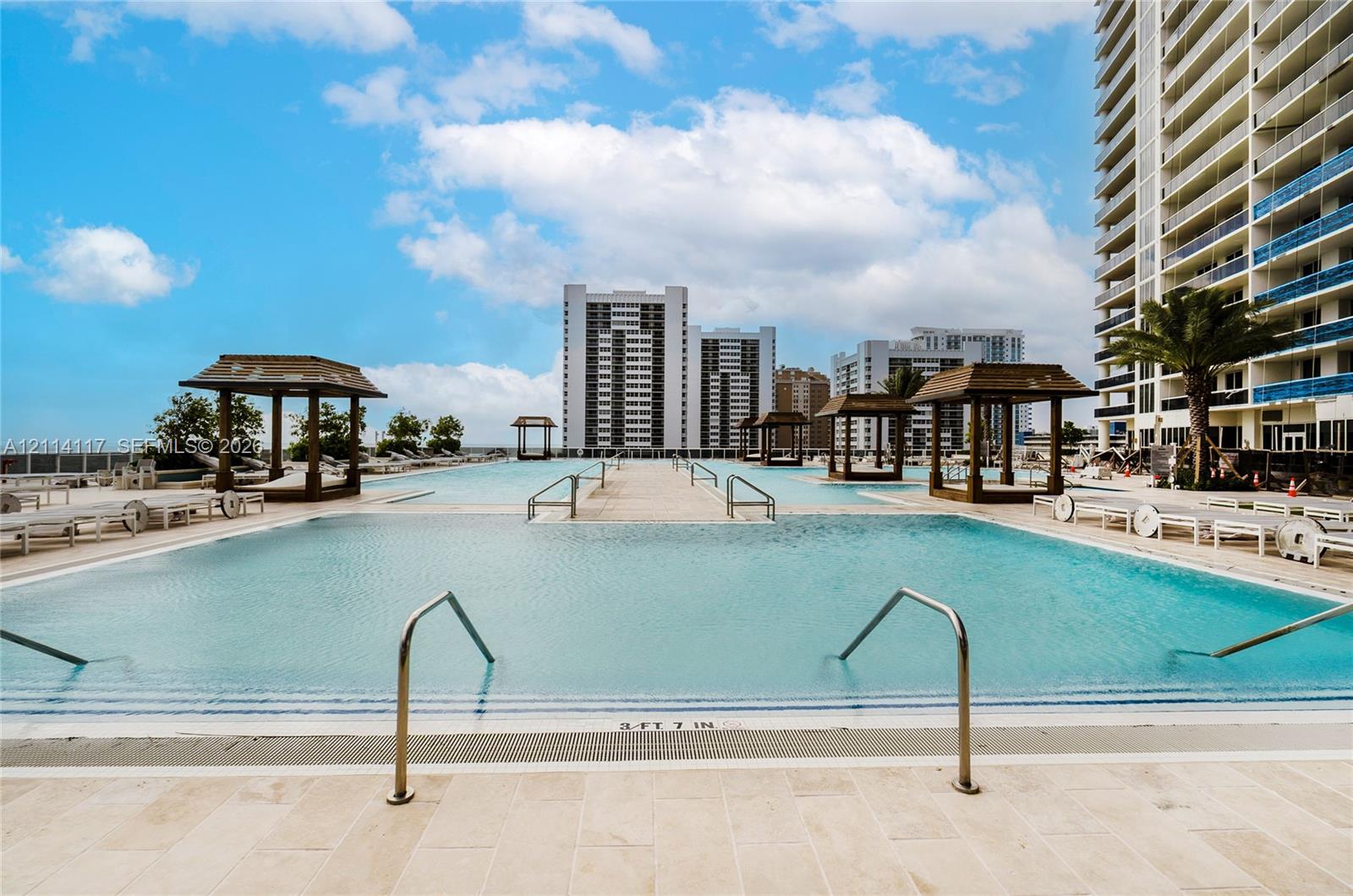 Large outdoor swimming pool with submerged entry steps, stainless steel handrails, tiled pool deck, and multiple wooden cabana structures