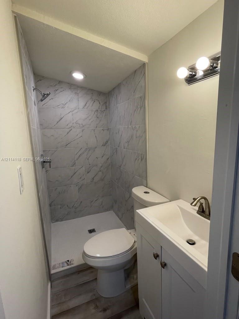 Bathroom featuring a shower stall with stone-patterned wall tile, white vanity cabinet with a integrated sink, polished metal hardware, wood-finish flooring, and wall-mounted vanity lighting