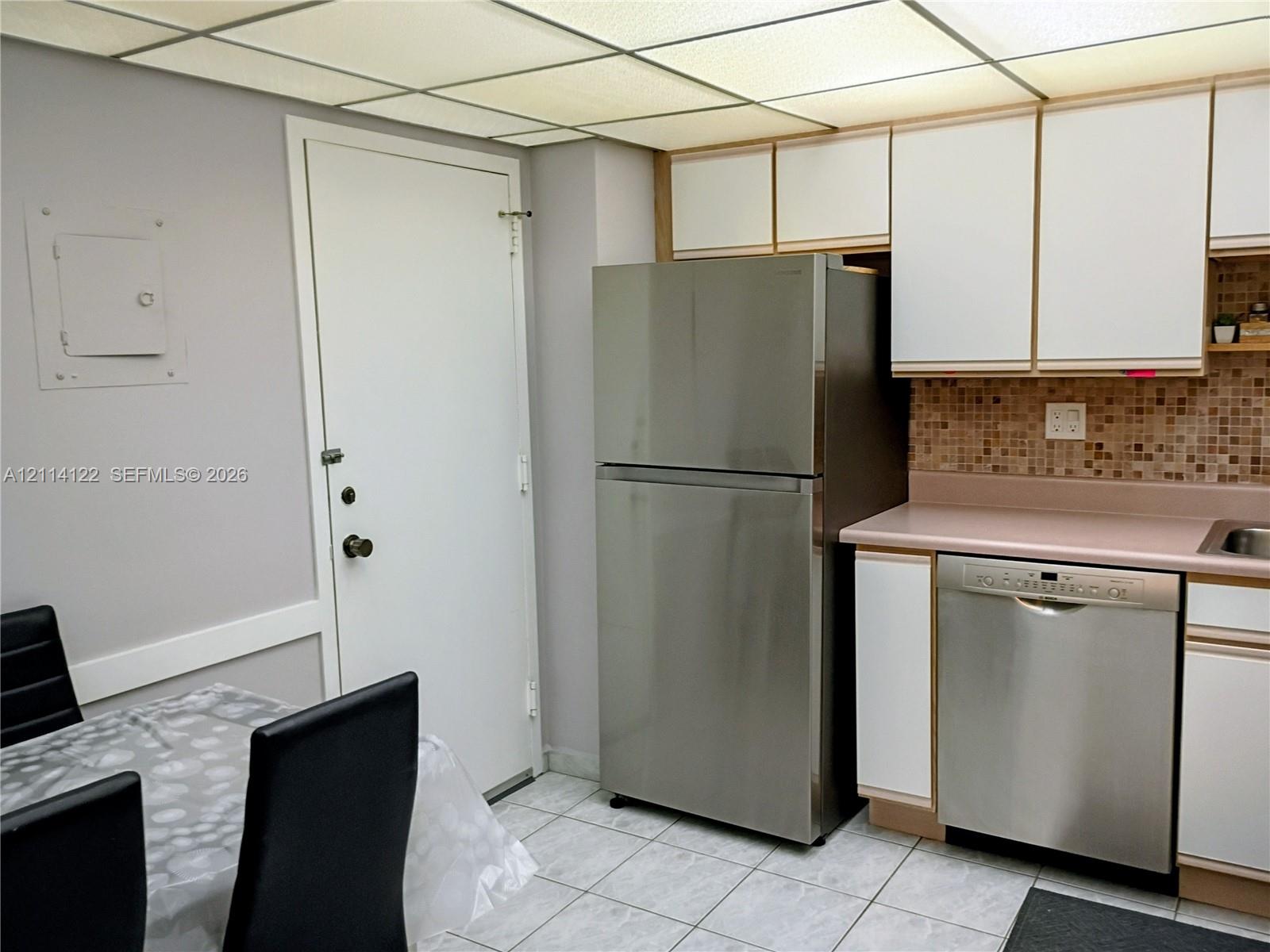 Kitchen area featuring stainless steel refrigerator and dishwasher, tiled backsplash, cabinetry with white finish, and tile flooring Kitchen area featuring stainless steel refrigerator and dishwasher, tiled backsplash, cabinetry with white finish, and tile flooring