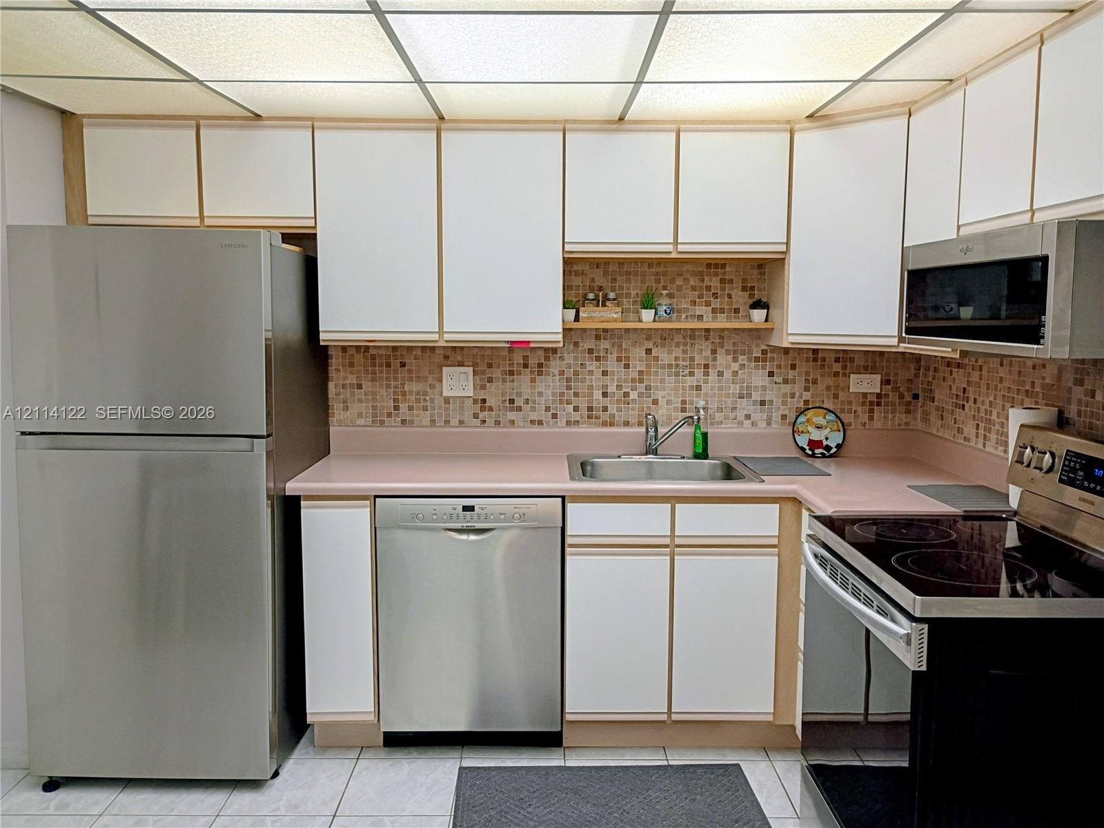 Kitchen featuring stainless steel appliances, flat-panel cabinetry, mosaic tile backsplash, solid surface countertops, and a grid-panel drop ceiling Kitchen featuring stainless steel appliances, flat-panel cabinetry, mosaic tile backsplash, solid surface countertops, and a grid-panel drop ceiling