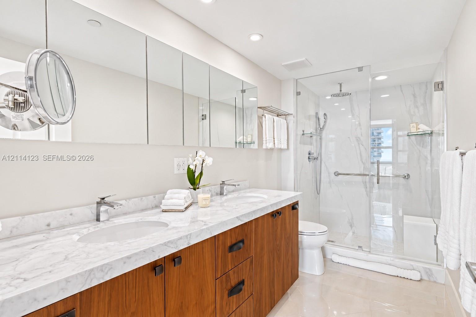 Double vanity featuring a light-toned stone countertop with dual undermount sinks, wood-finish cabinetry, floor-to-ceiling mirrored medicine cabinets, and a glass-enclosed shower with tiled walls and a built-in bench