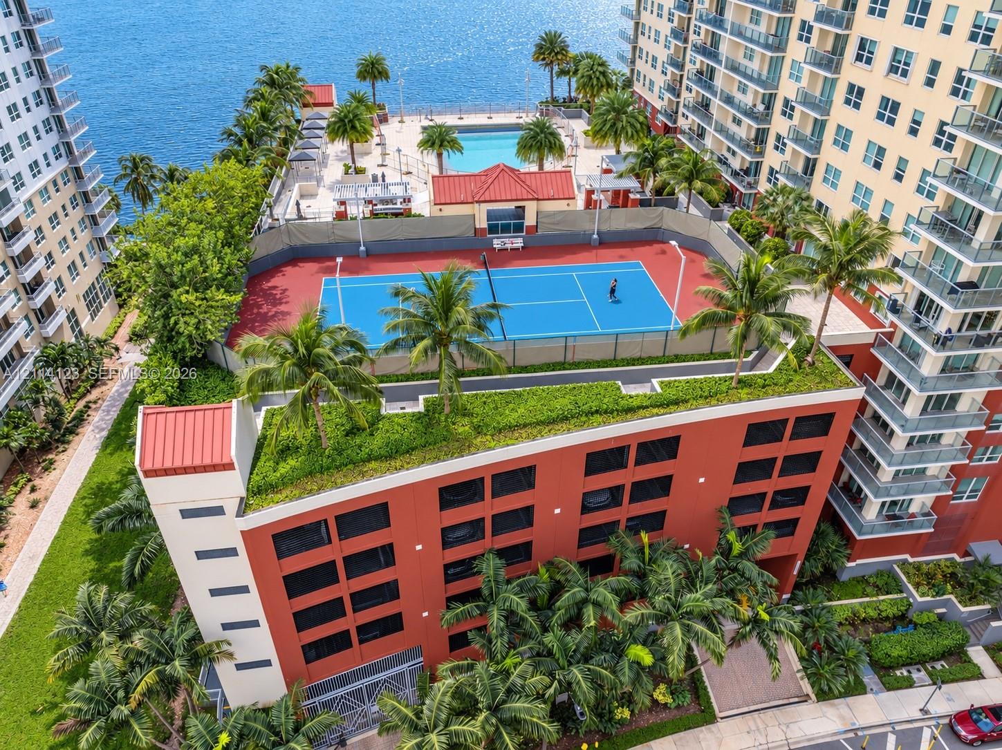 Rooftop amenities featuring a tennis court, swimming pool, mature palm trees, and waterfront perimeter
