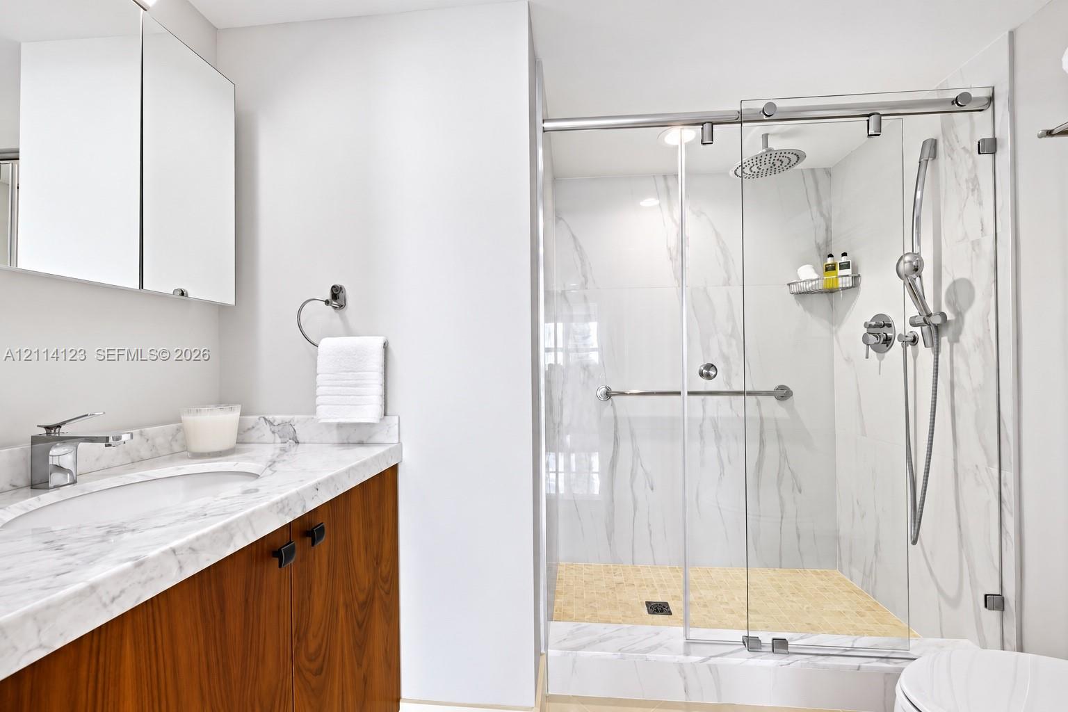 Bathroom featuring a wood-finish vanity with a stone-top integrated sink, stone-surround shower with a glass sliding door, rainfall showerhead, and a mirrored medicine cabinet