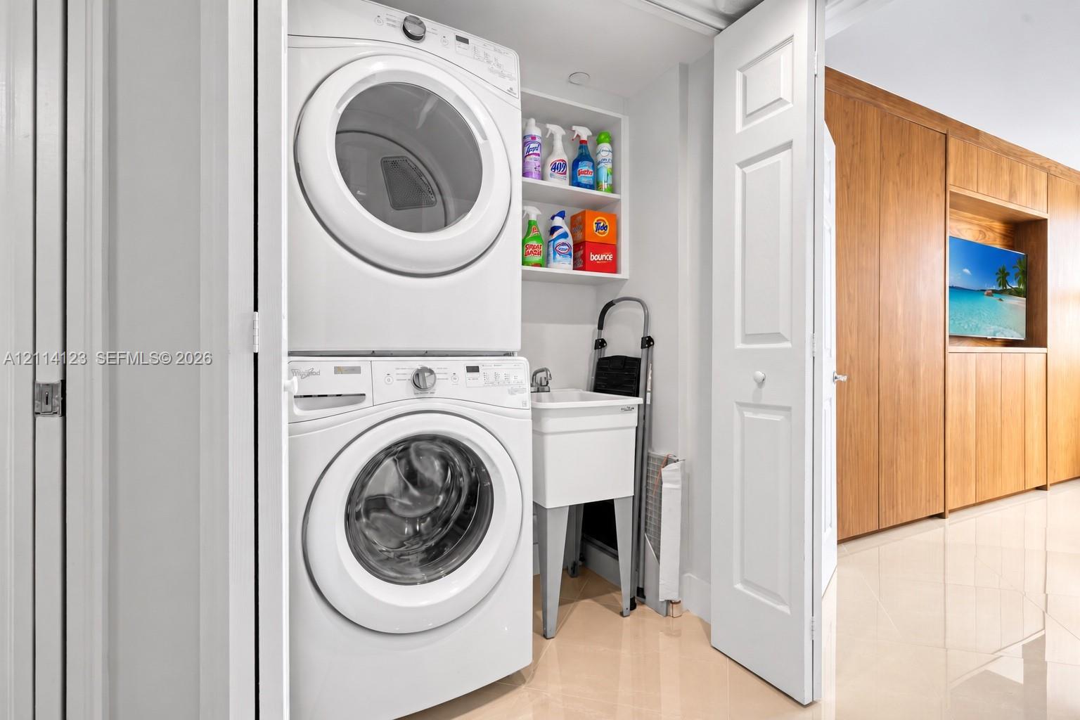 Laundry closet featuring a stacked front-load washer and dryer, utility sink, overhead shelving, and floor-to-ceiling wood-finish cabinetry