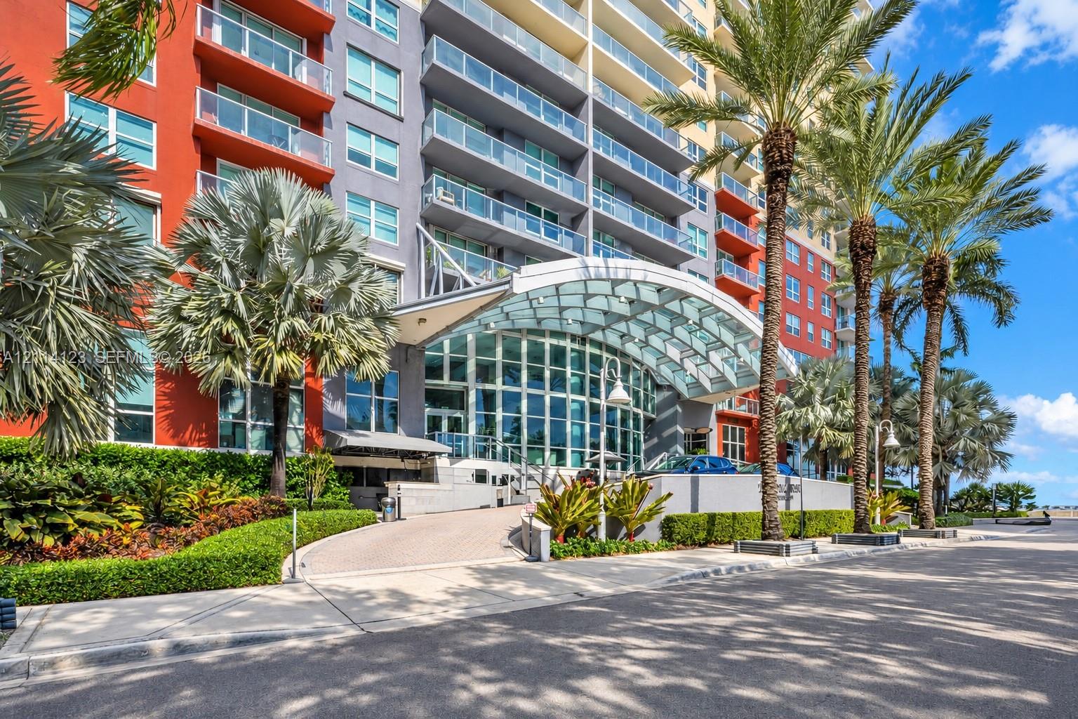 High-rise exterior featuring a glass-canopied entryway, floor-to-ceiling windows, private balconies with glass railings, and mature palm tree landscaping