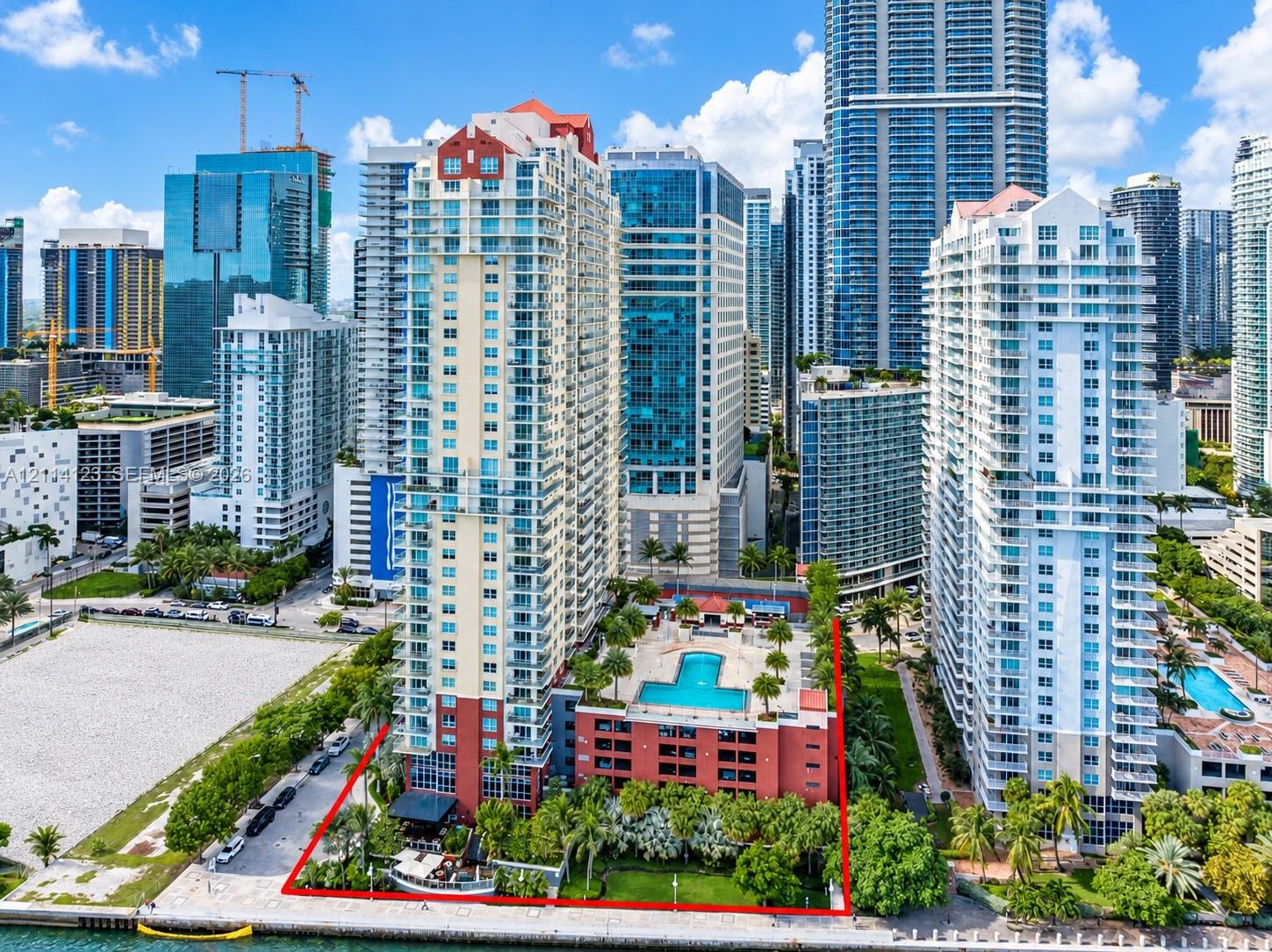 High-rise residential complex featuring an elevated pool deck, mature landscaping, ground-level waterfront promenade, and multi-story structural parking