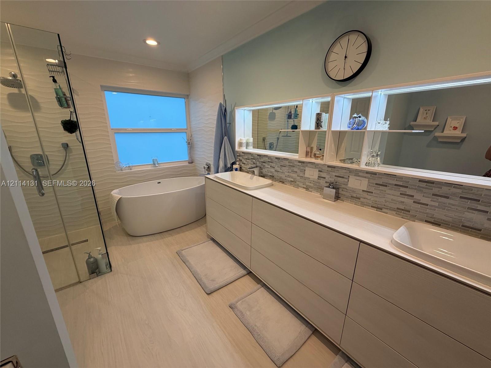 Double vanity with rectangular vessel sinks, freestanding soaking tub, glass-enclosed walk-in shower, horizontal tile backsplash, and light-tone cabinetry