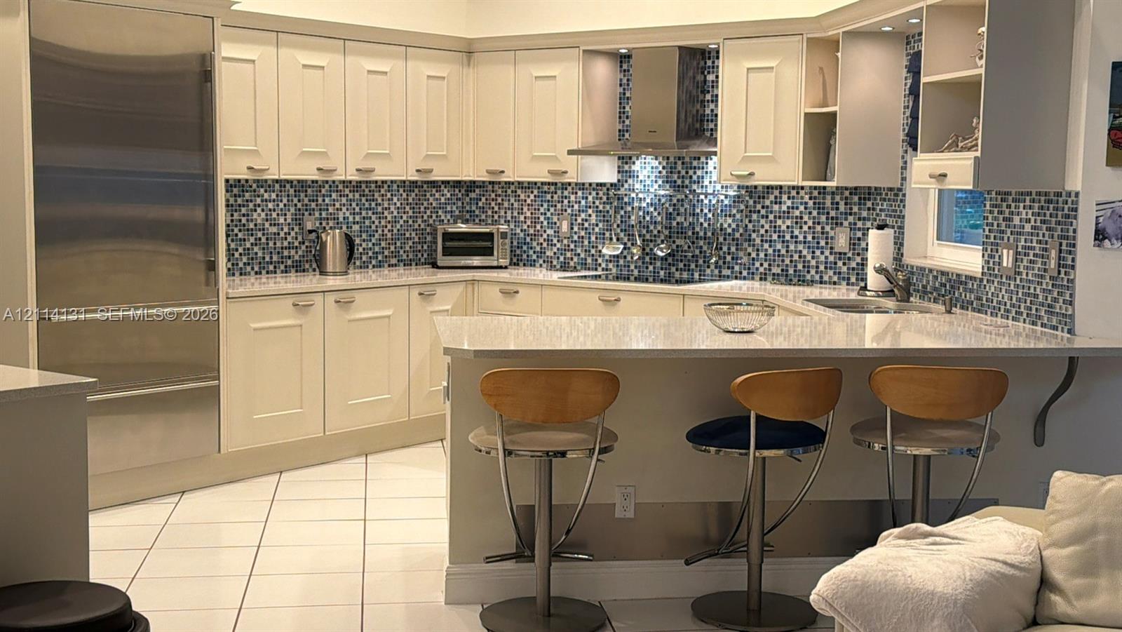 Kitchen featuring cream cabinetry, stone-finish countertops, full-height blue glass tile backsplash, stainless steel refrigerator, and stainless steel range hood
