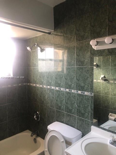 Bathroom featuring full-height green ceramic wall tiling with a decorative mosaic border, a built-in bathtub, a white vanity with an integrated sink, and a dual-bulb vanity light fixture Bathroom featuring full-height green ceramic wall tiling with a decorative mosaic border, a built-in bathtub, a white vanity with an integrated sink, and a dual-bulb vanity light fixture