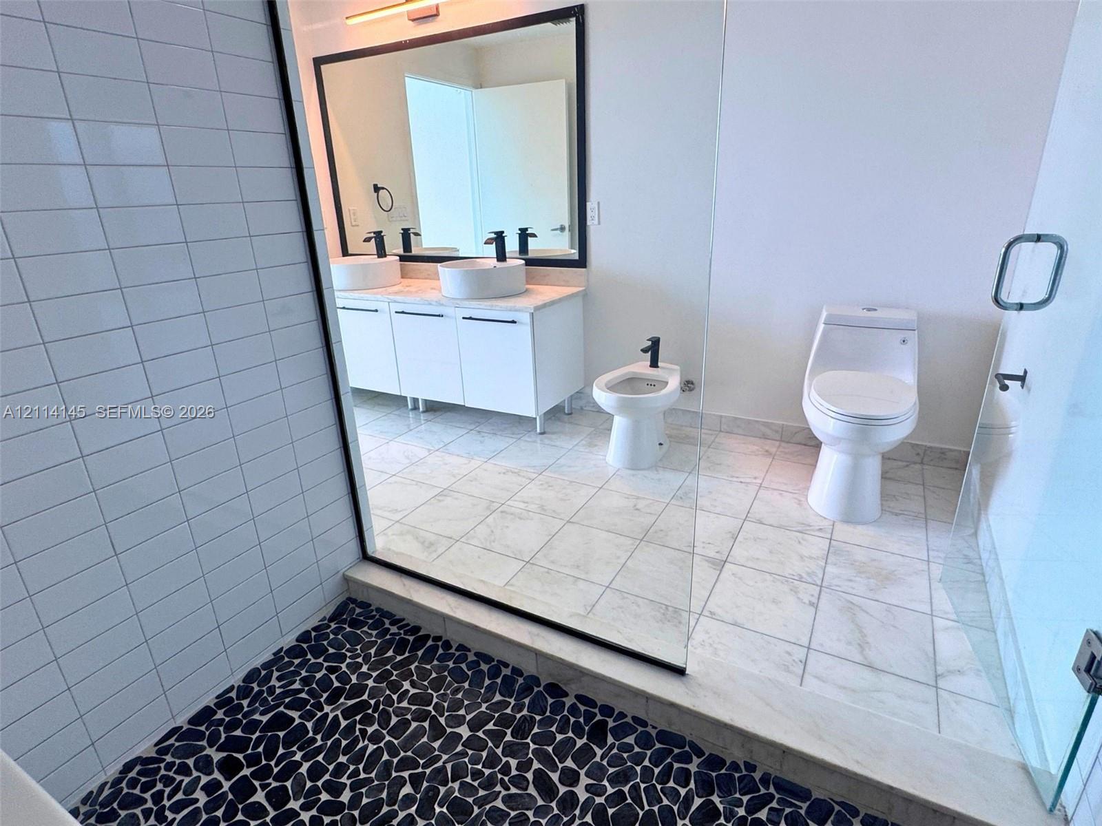 Bathroom featuring a dual-sink vanity with matte black faucets, a stone-tile shower floor, glass shower enclosure, and neutral stone-finish flooring