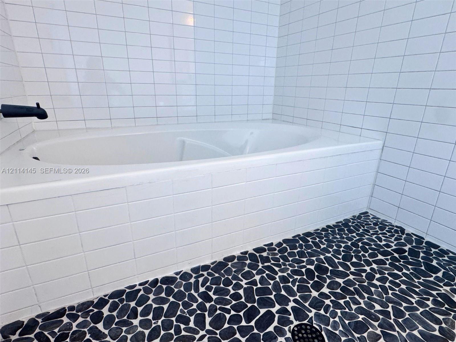 Built-in soaking tub with white square tile surround, matte black wall-mounted faucet, and natural stone pebble flooring