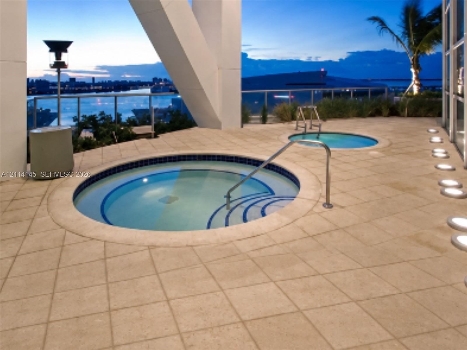 Elevated terrace featuring dual circular spas with tile lining, stone-finish paving, glass balustrades, and integrated ground-level lighting
