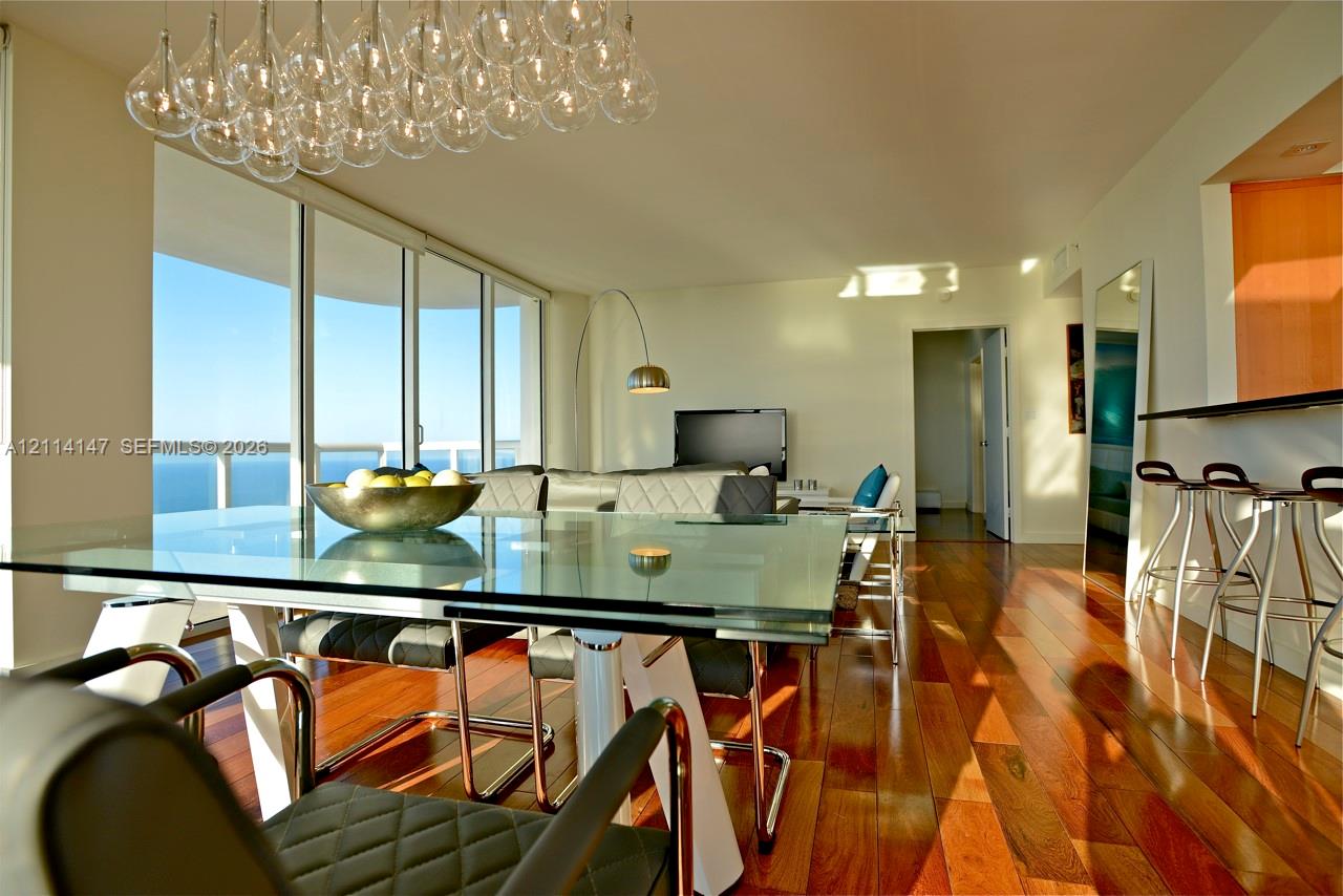 Floor-to-ceiling glass windows, wide-plank wood-finish flooring, custom multi-pendant chandelier, and a built-in breakfast bar