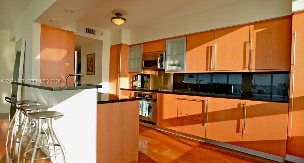 Kitchen featuring wood cabinetry, stainless steel appliances, dark stone-finish countertops, an integrated breakfast bar, and wood-finish flooring