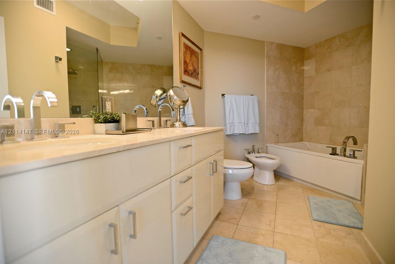 Double vanity with stone countertop, undermount sinks, and polished chrome faucets, featuring a full-width mirror, tile flooring, and a tiled shower-tub combination