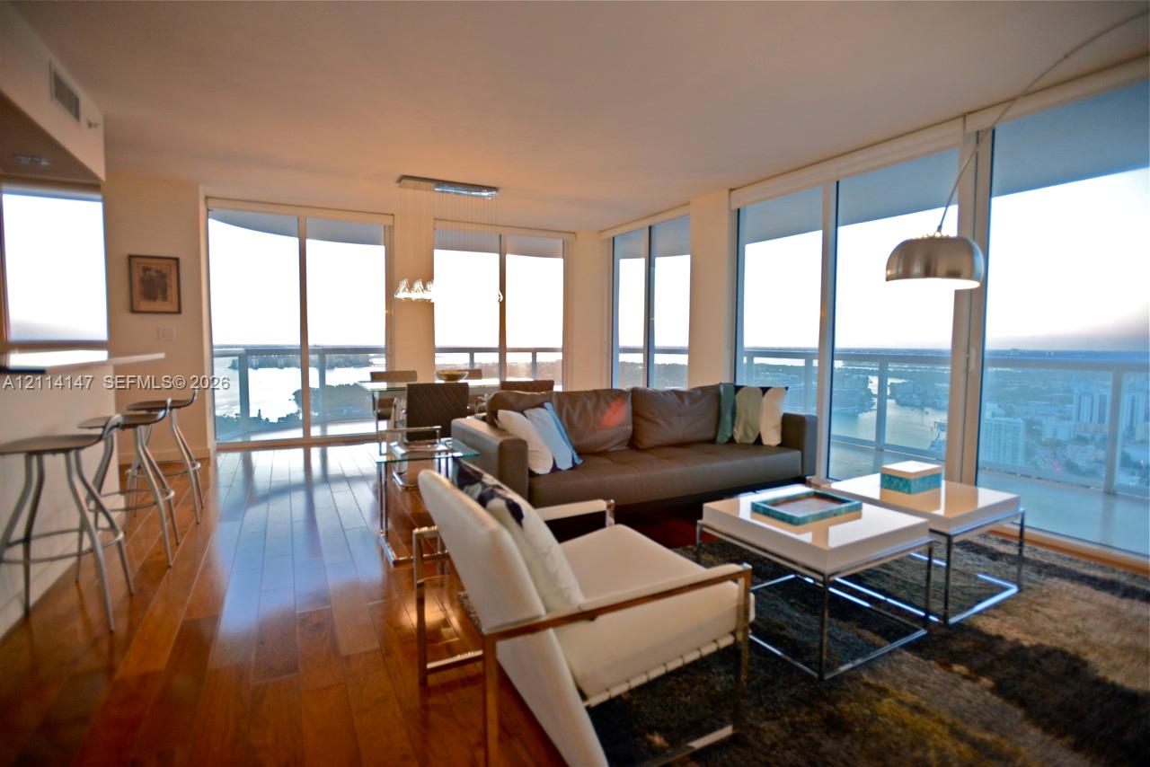 Open-concept interior featuring floor-to-ceiling glass walls, expansive water scenery, wood-finish flooring, and a built-in kitchen breakfast bar