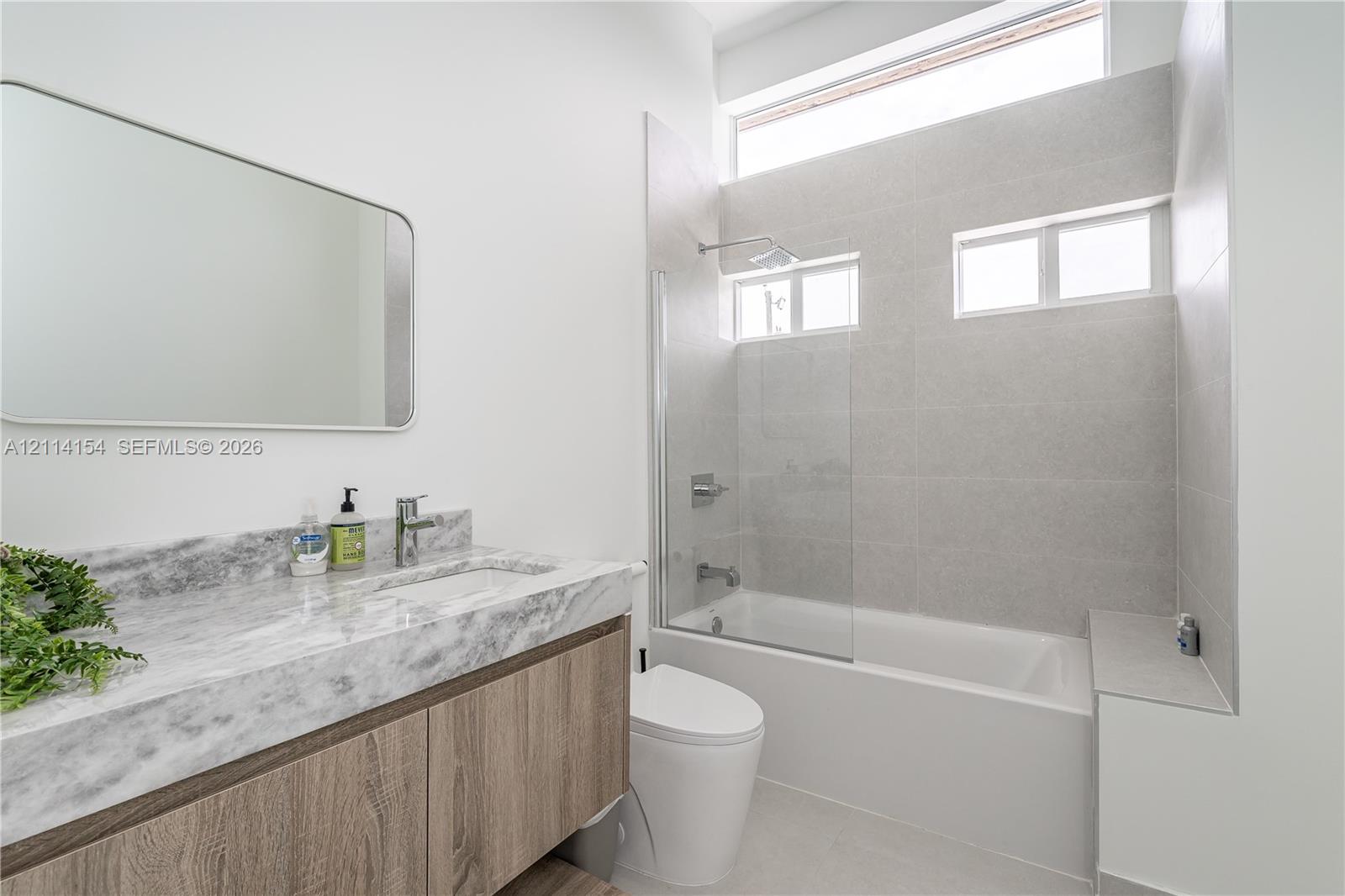 Floating wood-finish vanity with stone-surface countertop, rectangular undermount sink, glass-enclosed tub shower with full-height tile surround, and clerestory window