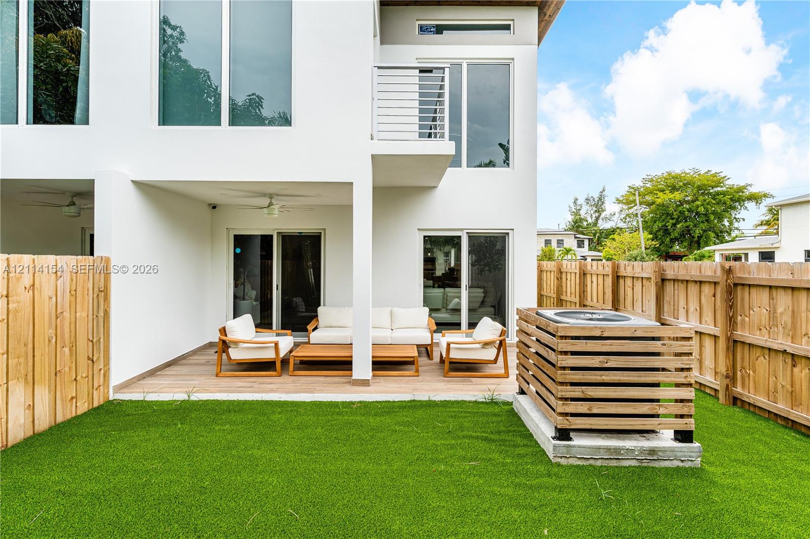 Fenced backyard featuring a covered patio with wood-finish decking, synthetic turf lawn, second-story balcony with metal railing, and horizontal slat privacy screen for exterior mechanical equipment
