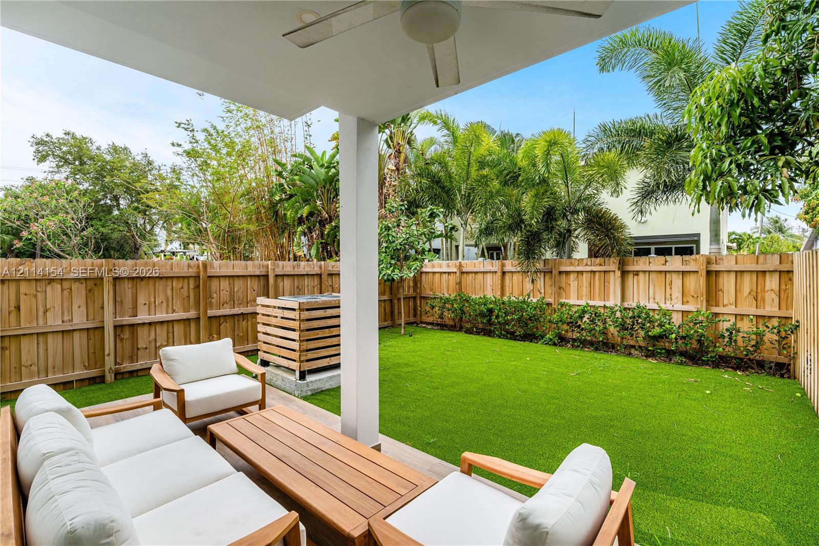 Covered patio featuring a ceiling fan, wood-style fencing, synthetic turf lawn, and exterior privacy landscaping
