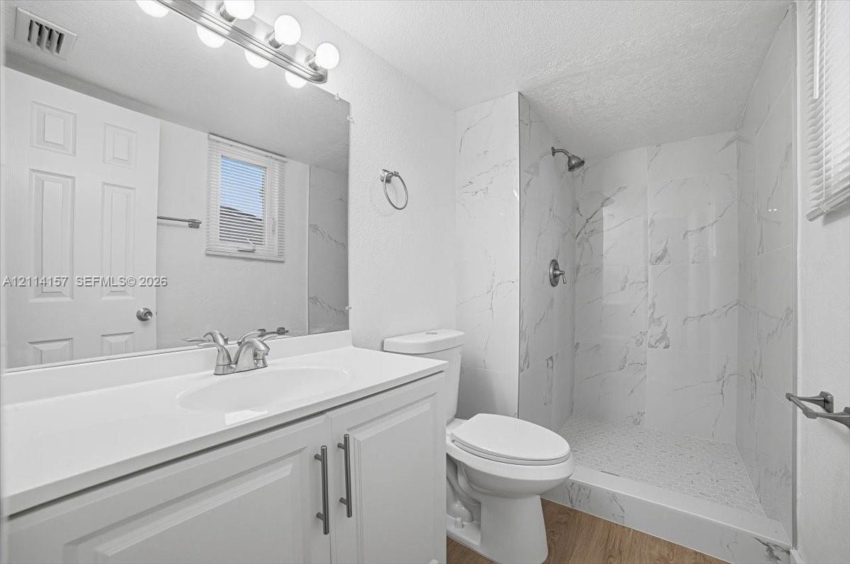 Bathroom featuring a white vanity with integrated sink, walk-in shower with floor-to-ceiling stone-pattern tile, polished chrome fixtures, and wood-finish flooring