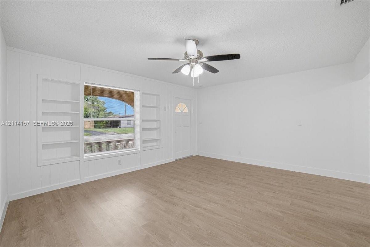 Interior space featuring wood-finish flooring, built-in shelving flanking a large window, a ceiling fan with integrated lighting, and a single-light panel entry door