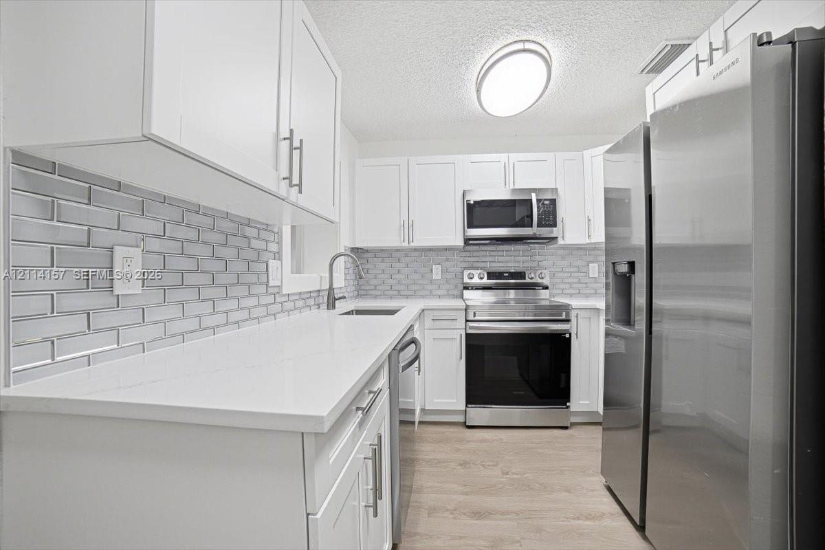 Kitchen featuring white shaker-style cabinetry, white solid-surface countertops, a gray subway tile backsplash, stainless steel appliances, and wood-finish flooring Kitchen featuring white shaker-style cabinetry, white solid-surface countertops, a gray subway tile backsplash, stainless steel appliances, and wood-finish flooring