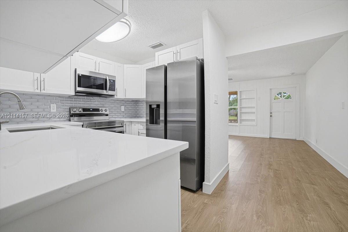 Kitchen featuring white shaker-style cabinetry, solid-surface countertops, a brick-patterned tile backsplash, stainless steel appliances, and wood-finish flooring Kitchen featuring white shaker-style cabinetry, solid-surface countertops, a brick-patterned tile backsplash, stainless steel appliances, and wood-finish flooring