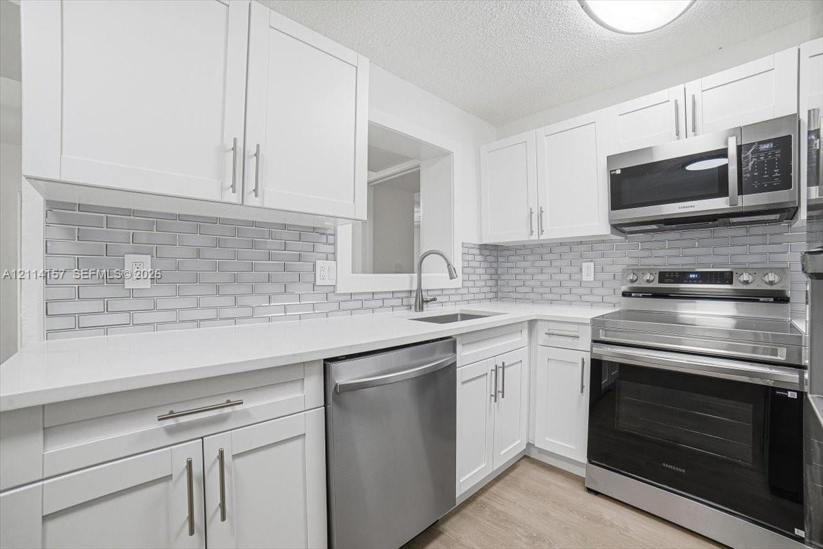 Kitchen featuring white shaker-style cabinetry, stainless steel appliances, white solid-surface countertops, light grey subway tile backsplash, and wood-finish flooring Kitchen featuring white shaker-style cabinetry, stainless steel appliances, white solid-surface countertops, light grey subway tile backsplash, and wood-finish flooring