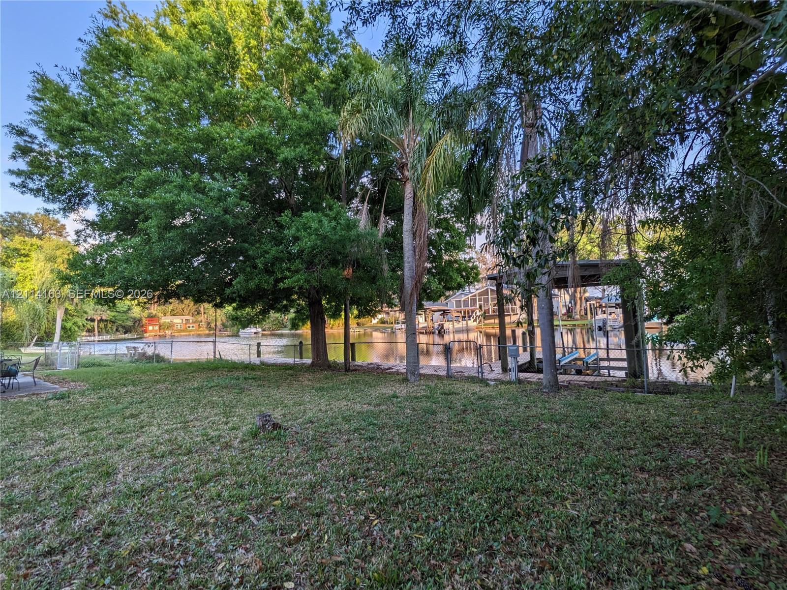 Waterfront grounds featuring mature canopy trees, a palm tree, a paved stone patio, and a private wooden boat dock