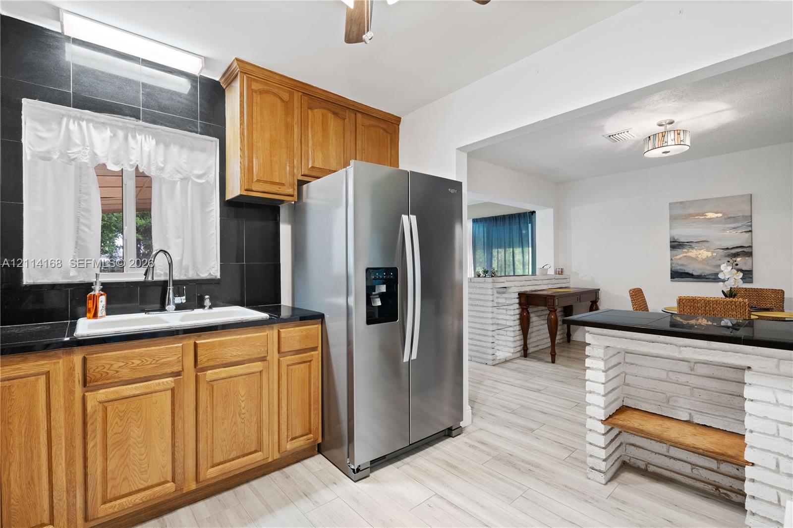 Kitchen featuring wood cabinetry, black tiled backsplash, stainless steel refrigerator, white dual-basin sink, and a white brick breakfast bar with a black countertop