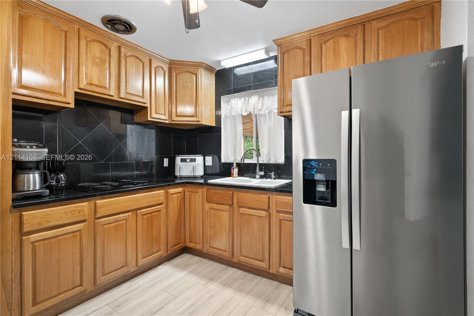 Kitchen featuring natural-finish wood cabinetry, black tile backsplash, stainless steel refrigerator, white double-basin sink, and wood-finish flooring