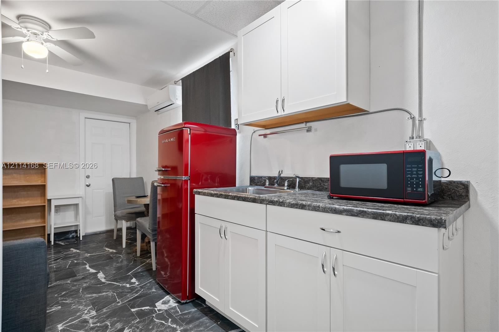 Kitchenette area featuring white shaker-style cabinetry, dark stone-finish countertops, stainless steel sink, wall-mounted ductless climate control unit